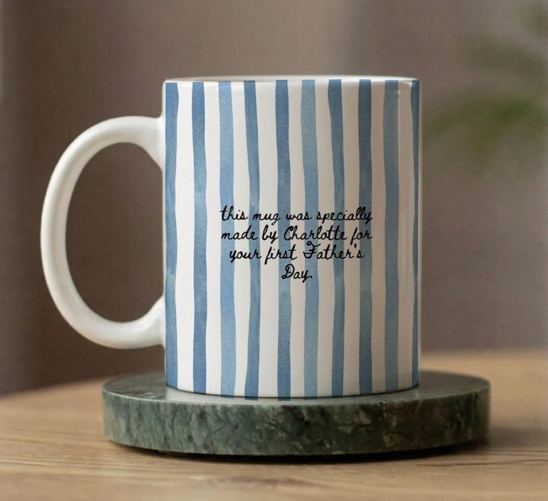 Op de afbeelding: Witte keramische mok met blauwe verticale strepen. De mok heeft een wit handvat en bevat de tekst: "this mug was specially made by Charlotte for your first Father's Day."