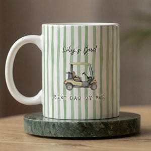 Puede incluir: Taza de cerámica blanca con rayas verticales verdes, con una ilustración de carrito de golf y el texto "Lily's Dad BEST DAD BY PAR". La taza está sobre un posavasos de mármol verde.