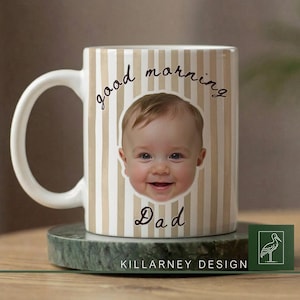 Op de afbeelding: Witte keramische mok met een beige en wit gestreept patroon. De mok toont het gezicht van een lachende baby en de woorden "good morning Dad" in zwart schrift. De mok staat op een groene marmeren onderzetter.