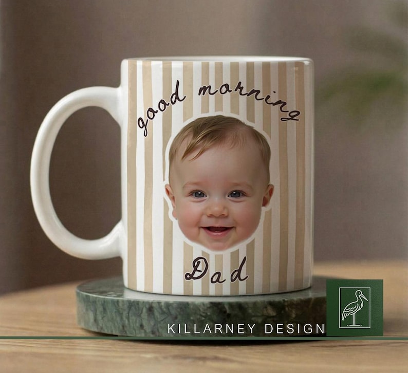 Op de afbeelding: Witte keramische mok met een beige en wit gestreept patroon. De mok toont het gezicht van een lachende baby en de woorden "good morning Dad" in zwart schrift. De mok staat op een groene marmeren onderzetter.