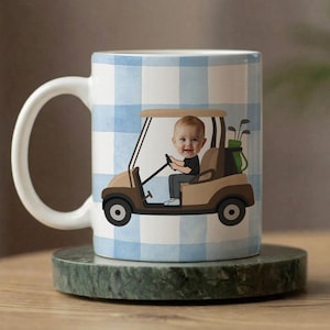 Puede incluir: Taza de cerámica blanca con un patrón de cuadros azules y blancos. La taza presenta una ilustración de dibujos animados de un bebé conduciendo un carrito de golf. El carrito de golf es de color beige y marrón, con palos de golf en la parte trasera.