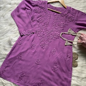 Pu&ograve; includere: Una tunica color lavanda con ricami floreali intricati. Il top a maniche a tre quarti presenta cuciture dettagliate intorno allo scollo, alle maniche e al corpo. La tunica &egrave; esposta su una gruccia di legno.