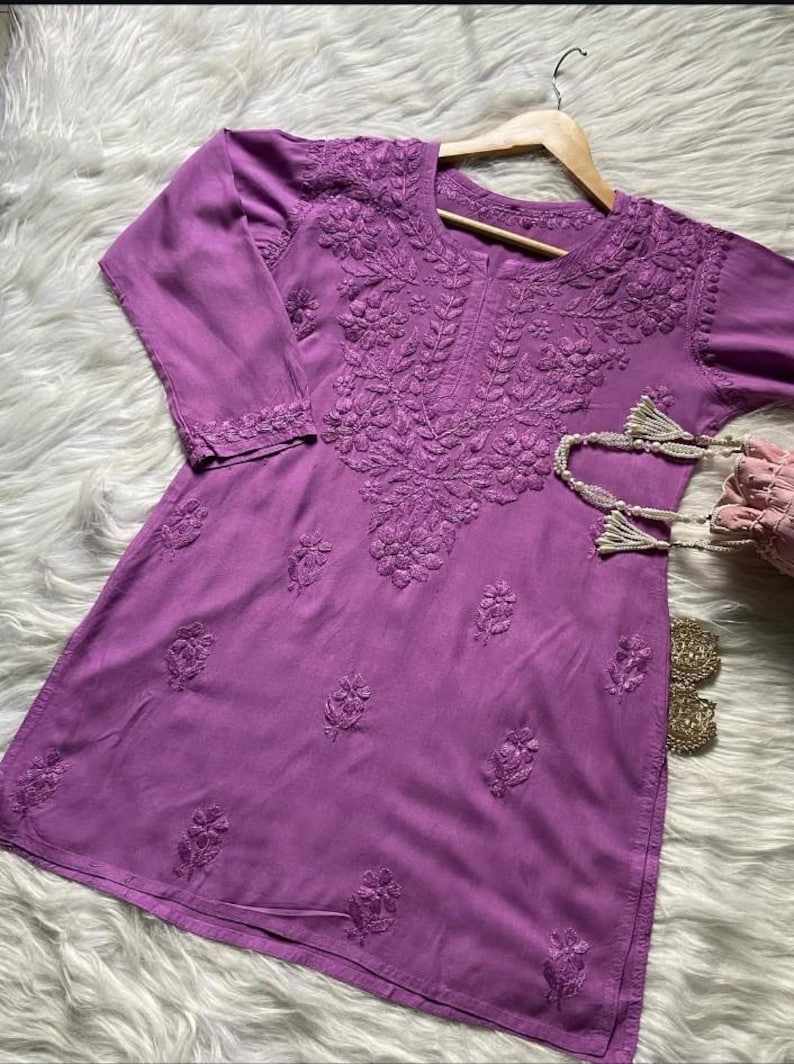 Pu&ograve; includere: Una tunica color lavanda con ricami floreali intricati. Il top a maniche a tre quarti presenta cuciture dettagliate intorno allo scollo, alle maniche e al corpo. La tunica &egrave; esposta su una gruccia di legno.