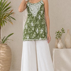 Op de afbeelding: Een vrouw draagt een groen geborduurd topje en een witte palazzo broek. De top heeft een bloemenpatroon en spaghettibandjes. De broek is wijd en wit. De outfit wordt gecompleteerd met witte sandalen. De achtergrond is neutraal.