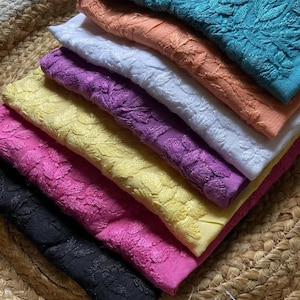 Pu&ograve; includere: Una pila di tessuti ricamati in vari colori, tra cui turchese, pesca, bianco, viola, giallo, rosa e nero. Il tessuto presenta un motivo floreale ed &egrave; ordinatamente disposto su un tappetino intrecciato. L'immagine mostra una variet&agrave; di tessuti.