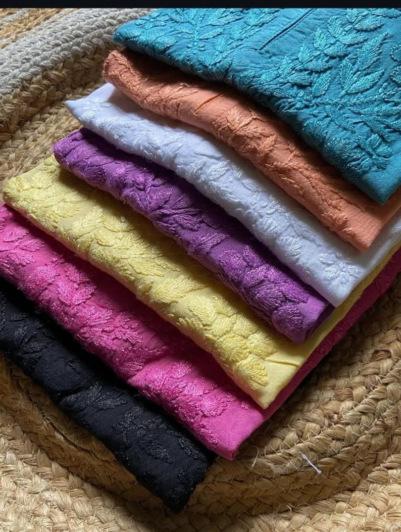 Pu&ograve; includere: Una pila di tessuti ricamati in vari colori, tra cui turchese, pesca, bianco, viola, giallo, rosa e nero. Il tessuto presenta un motivo floreale ed &egrave; ordinatamente disposto su un tappetino intrecciato. L'immagine mostra una variet&agrave; di tessuti.