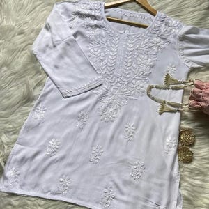 Pu&ograve; includere: Tunica bianca con ricami floreali intricati. Il top a maniche lunghe presenta cuciture dettagliate intorno allo scollo, alle maniche e al corpo. L'indumento &egrave; esposto su un'appendiabiti di legno su uno sfondo strutturato.