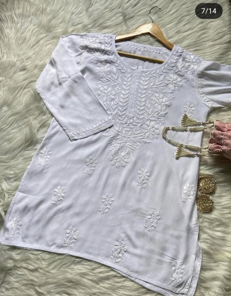 Pu&ograve; includere: Tunica bianca con ricami floreali intricati. Il top a maniche lunghe presenta cuciture dettagliate intorno allo scollo, alle maniche e al corpo. L'indumento &egrave; esposto su un'appendiabiti di legno su uno sfondo strutturato.