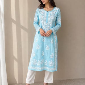 May include: A light blue long-sleeved tunic featuring white floral embroidery along the neckline, sleeves, and hem, paired with plain white straight-leg trousers. The tunic has a round neckline with a small slit and side vents at the hem.