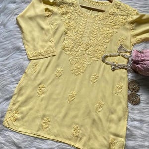 Pu&ograve; includere: Una tunica giallo pallido con ricami floreali intricati. La tunica presenta uno scollo tondo e maniche a tre quarti. Il ricamo &egrave; concentrato intorno allo scollo e sparso sul davanti. Sono visibili una borsetta rosa e orecchini dorati.