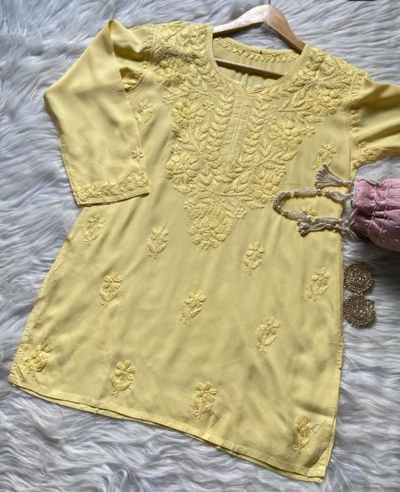 Pu&ograve; includere: Una tunica giallo pallido con ricami floreali intricati. La tunica presenta uno scollo tondo e maniche a tre quarti. Il ricamo &egrave; concentrato intorno allo scollo e sparso sul davanti. Sono visibili una borsetta rosa e orecchini dorati.