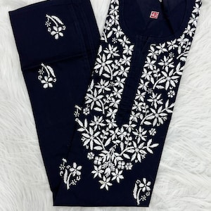 May include: Navy blue embroidered tunic and pants set. The tunic features a floral design in white thread around the neckline and down the front. The pants have matching floral embroidery. The set is displayed on a white, fluffy surface.