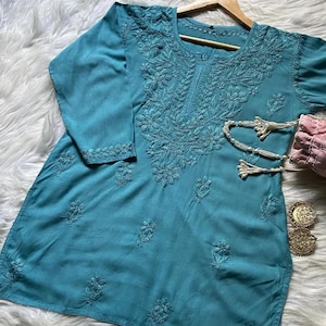 以下が含まれることがあります： 精巧な花の刺繍が施されたターコイズ色のチュニック。長袖のトップスは木製のハンガーに掛けられています。ビーズのネックレス、ピンクの財布、金のイヤリングが見えます。