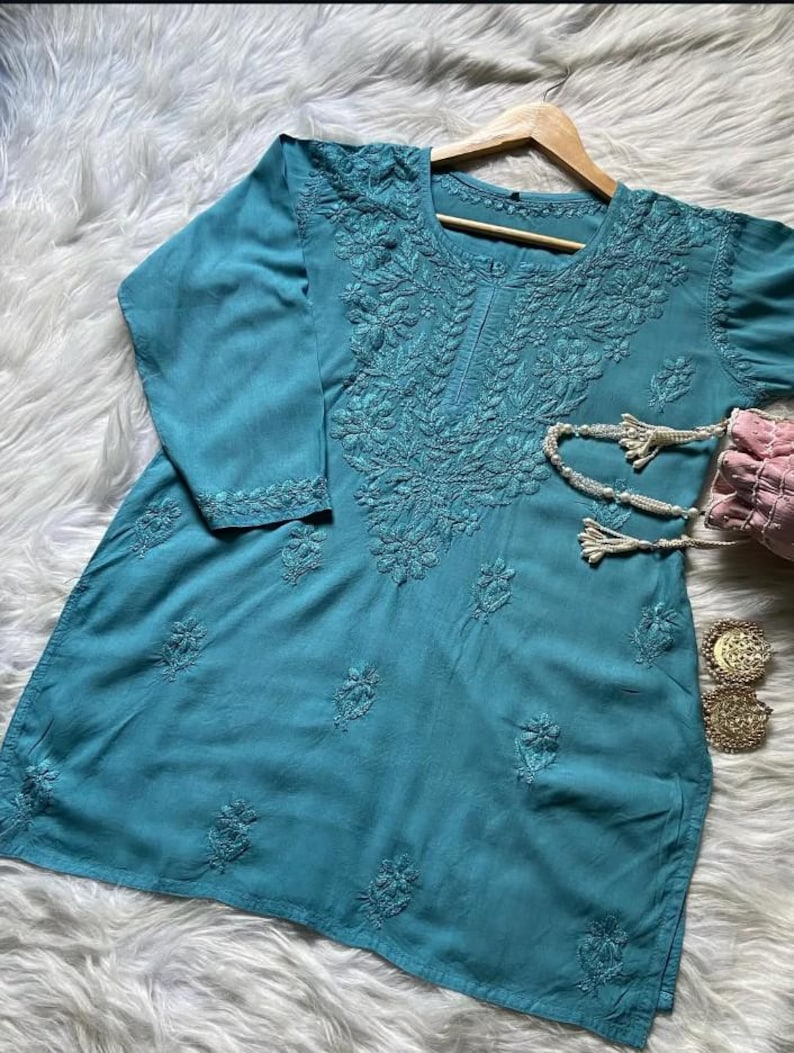Pu&ograve; includere: Una tunica turchese con ricami floreali intricati. La parte superiore a maniche lunghe &egrave; esposta su un appendiabiti in legno. Sono visibili una collana di perline, una borsetta rosa e orecchini dorati.