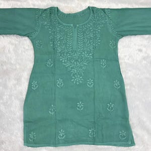 Könnte beinhalten: Eine meergrüne Tunika mit langen Ärmeln, einem Rundhalsausschnitt mit einem kleinen Schlitz in der Mitte sowie tonalen Blumenstickereien, die sich im Brustbereich konzentrieren und über den unteren Teil des Stoffes verteilt sind.