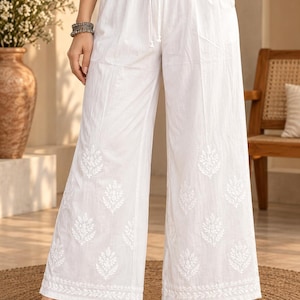 May include: White wide-leg trousers featuring an elasticized drawstring waistband and white floral embroidery patterns distributed across the lower portion of the legs, finished with a detailed embroidered border along the bottom hemline.