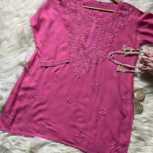 Pu&ograve; includere: Una tunica magenta con ricami floreali intricati intorno allo scollo, alle maniche e al corpo. Il top a maniche lunghe &egrave; esposto su una gruccia di legno. Sono visibili una piccola borsa rosa e orecchini color oro.