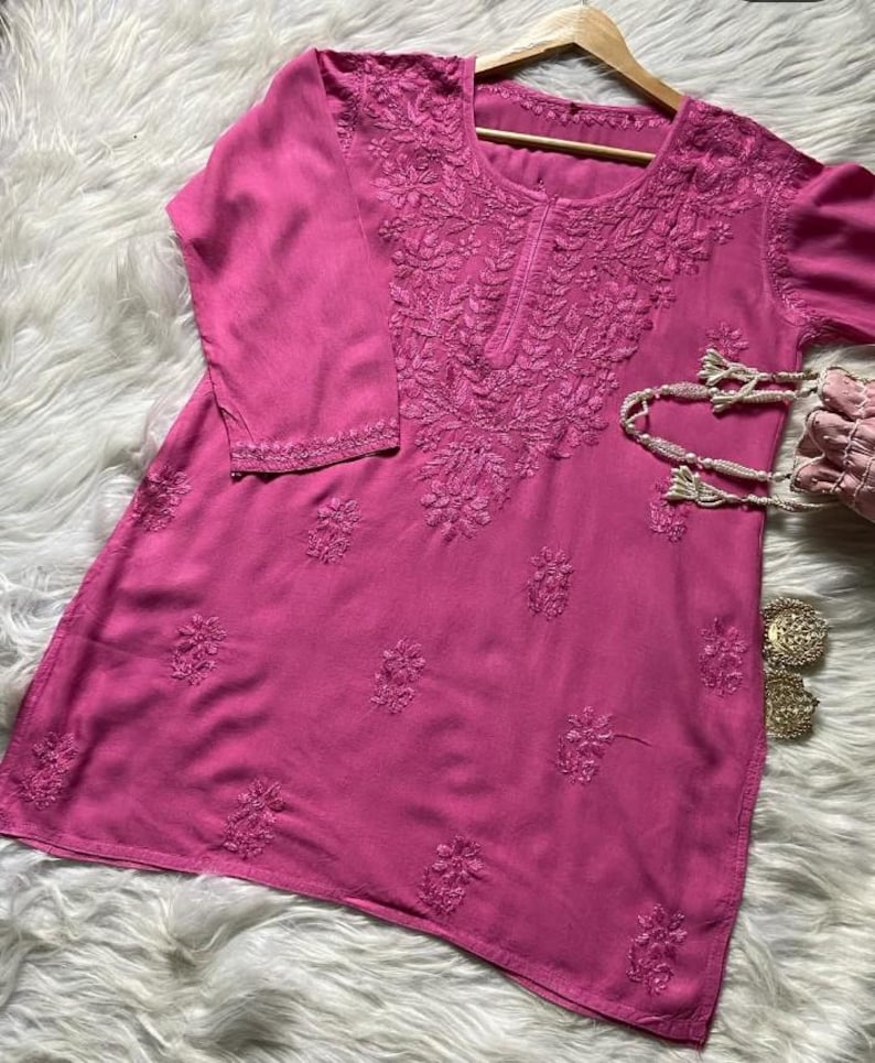 Pu&ograve; includere: Una tunica magenta con ricami floreali intricati intorno allo scollo, alle maniche e al corpo. Il top a maniche lunghe &egrave; esposto su una gruccia di legno. Sono visibili una piccola borsa rosa e orecchini color oro.