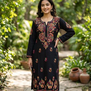 Könnte beinhalten: Eine schwarze Tunika mit aufwendiger, farbenfroher Blumenstickerei. Die Stickerei weist Farbtöne von Rot, Orange, Gelb und Grün auf. Die Tunika hat einen V-Ausschnitt und wird mit einer schwarzen Hose getragen. Die Frau lächelt und trägt goldene Ohrringe.