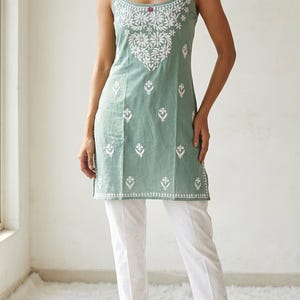 May include: A sage green sleeveless tunic featuring white floral embroidery on the bodice and hem, paired with plain white trousers. The tunic has thin spaghetti straps and a scoop neckline, with a small red detail at the center of the chest.