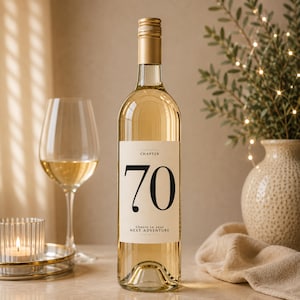 May include: A clear glass wine bottle with a gold cap and a cream-colored label. The label features the word CHAPTER, the large black number 70, and the text Cheers to your NEXT ADVENTURE. The bottle contains a light-colored liquid.