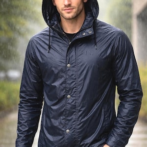 Può includere: Giacca antipioggia blu navy con cappuccio e chiusura con bottoni a pressione. La giacca ha un cappuccio con coulisse e maniche lunghe. La persona nell'immagine indossa la giacca sotto la pioggia.