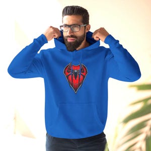 May include: A royal blue hoodie featuring a red and black spider emblem on the chest. The hoodie has a drawstring hood and a front pocket. The design is a graphic representation of a spider.