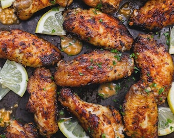 May include: An overhead shot of a tray filled with cooked chicken wings. The wings are golden brown with a slightly charred appearance, garnished with chopped parsley and lemon wedges. A small bowl of dipping sauce is visible.