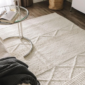 以下が含まれることがあります： クリーム色の、幾何学模様のテクスチャード加工されたラグ。ラグは、盛り上がった、ボブルのような質感と線状のデザインを組み合わせています。ニュートラルな色と触覚的な表面が、リビングルームやベッドルームに適した、居心地の良い魅力的な美学を作り出しています。