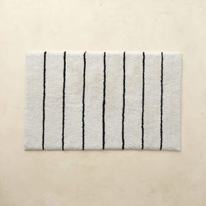 May include: A rectangular bath mat featuring a textured off-white surface with seven vertical black stripes. The mat has a soft, pile-like appearance and is oriented horizontally, showcasing a consistent pattern of thin, dark lines across the light fabric.