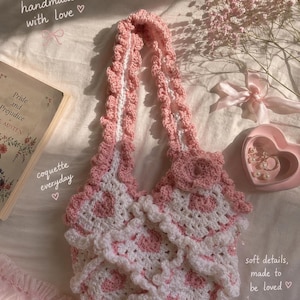 May include: A handmade crochet bag featuring a white and pink heart pattern with ruffled edges and a single strap. The bag is adorned with a crocheted flower accent. Text on the image reads: handmade with love, coquette everyday, soft details, made to be loved.