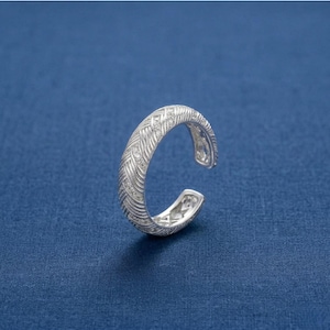 May include: A silver open cuff ring with a woven pattern and small, sparkling details. The ring is displayed on a blue backdrop, highlighting its detailed design and elegant style.