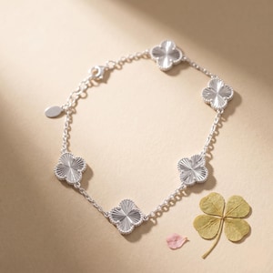 May include: A silver bracelet featuring five clover-shaped charms. The charms have a textured, radial design. The bracelet is on a neutral background with a four-leaf clover and a pink petal.