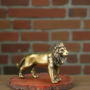 May include: A golden lion figurine, standing on a reddish-brown, circular wooden base. The lion has a detailed mane and is posed in a regal stance. The background features a brick wall.