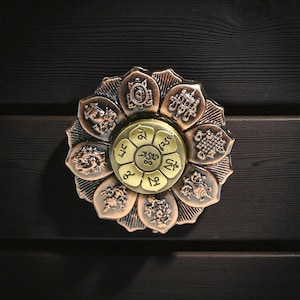 Può includere: Un oggetto decorativo a forma di loto color bronzo, con un disco centrale dorato. I petali presentano simboli e disegni in rilievo. L'oggetto è posto su uno sfondo scuro con venature del legno, creando un contrasto di trame e colori.