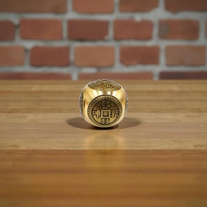 May include: A gold-colored, cube-shaped object with engraved Chinese characters. The cube is resting on a wooden surface, with a blurred brick wall in the background. The cube has a metallic sheen and appears to be made of metal.