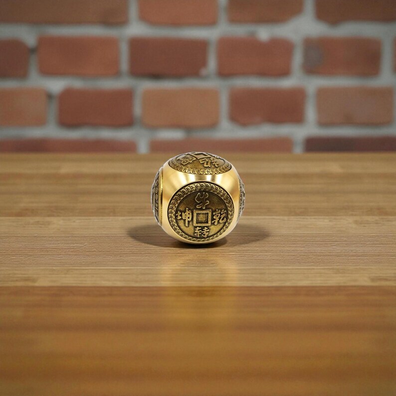 May include: A gold-colored, cube-shaped object with engraved Chinese characters. The cube is resting on a wooden surface, with a blurred brick wall in the background. The cube has a metallic sheen and appears to be made of metal.