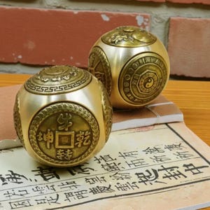 May include: Two brass-colored, spherical objects with intricate designs and Chinese characters. One sphere features a coin-like emblem with characters, while the other displays a Yin-Yang symbol. The objects rest on a paper with Asian script.