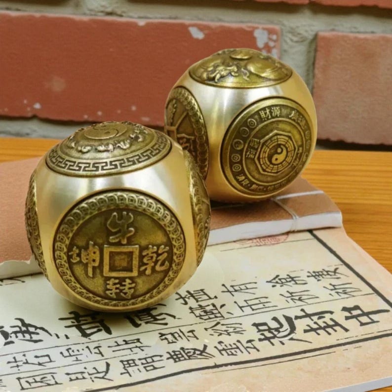 May include: Two brass-colored, spherical objects with intricate designs and Chinese characters. One sphere features a coin-like emblem with characters, while the other displays a Yin-Yang symbol. The objects rest on a paper with Asian script.