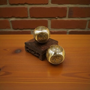 May include: Two gold-colored metal balls with intricate embossed designs, one resting on a small, dark wooden stand. The balls have a spherical shape with detailed patterns and symbols. The background features a wooden surface and a brick wall.