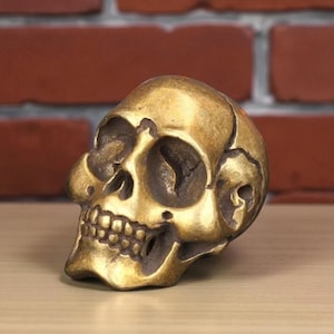 Op de afbeelding: Een messing schedelsculptuur met gedetailleerde kenmerken, waaronder oogkassen, neusholte en tanden. De schedel heeft een verweerde, antieke uitstraling, met zichtbare scheuren en onvolkomenheden. De achtergrond is een bakstenen muur en een houten oppervlak.