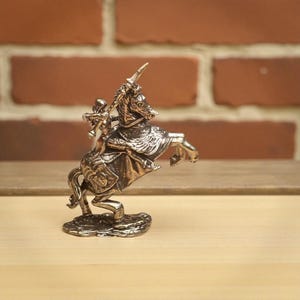 May include: A bronze-colored figurine of a knight on horseback, rearing up. The knight is holding a sword, and the horse is detailed with ornate armor. The figurine is mounted on a textured base, showcasing a medieval theme.