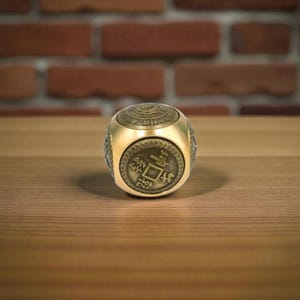 May include: A gold-colored, cube-shaped ring with intricate detailing. The ring features a coin-like design on its face and a patterned top. The ring is set against a wooden surface with a brick background.