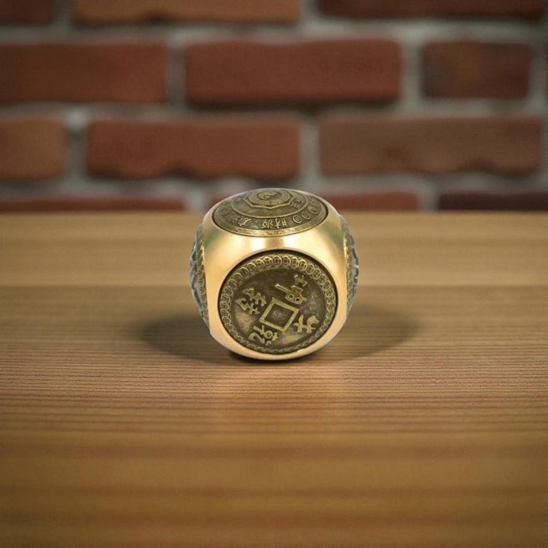 May include: A gold-colored, cube-shaped ring with intricate detailing. The ring features a coin-like design on its face and a patterned top. The ring is set against a wooden surface with a brick background.
