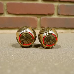 May include: Two gold-colored metal spheres with intricate engraved designs. Each sphere features a central circular design with Chinese characters and a red border. The spheres are displayed on a neutral surface against a brick background.