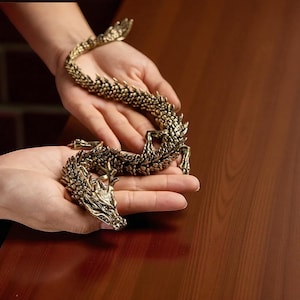 May include: A detailed, golden dragon figurine with intricate scales and features. The dragon is posed in a curved, serpentine shape, with its head and tail raised. The figurine is held in a person's hands, showcasing its size and detail.