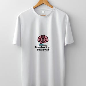 May include: White t-shirt with a crew neck and short sleeves. The shirt features a graphic of a pink brain with a loading bar and the text "Brain Loading... Please Wait". The design is centered on the front of the shirt.