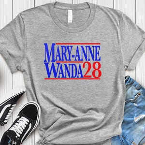 Pode incluir: Uma t-shirt cinza mesclada de manga curta com o texto azul MARY-ANNE WANDA e o número vermelho 28, acentuado por linhas vermelhas horizontais acima e abaixo do texto, exibida ao lado de ténis pretos e brancos e calções de ganga azuis.