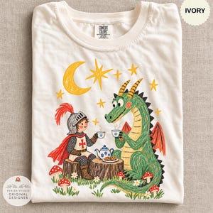 Puede incluir: Una camiseta de color marfil con una ilustración gráfica de un caballero con armadura y un dragón verde sentados en troncos de árboles, bebiendo té con galletas. El diseño incluye una luna creciente amarilla, estrellas y setas rojas.