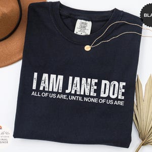 Op de afbeelding: Een zwart t-shirt met korte mouwen en de witte, verweerde tekst I AM JANE DOE ALL OF US ARE, UNTIL NONE OF US ARE. Het shirt heeft een Comfort Colors merklabel bij de hals en wordt vergezeld door een dunne goudkleurige ketting met een kleine ronde hanger.